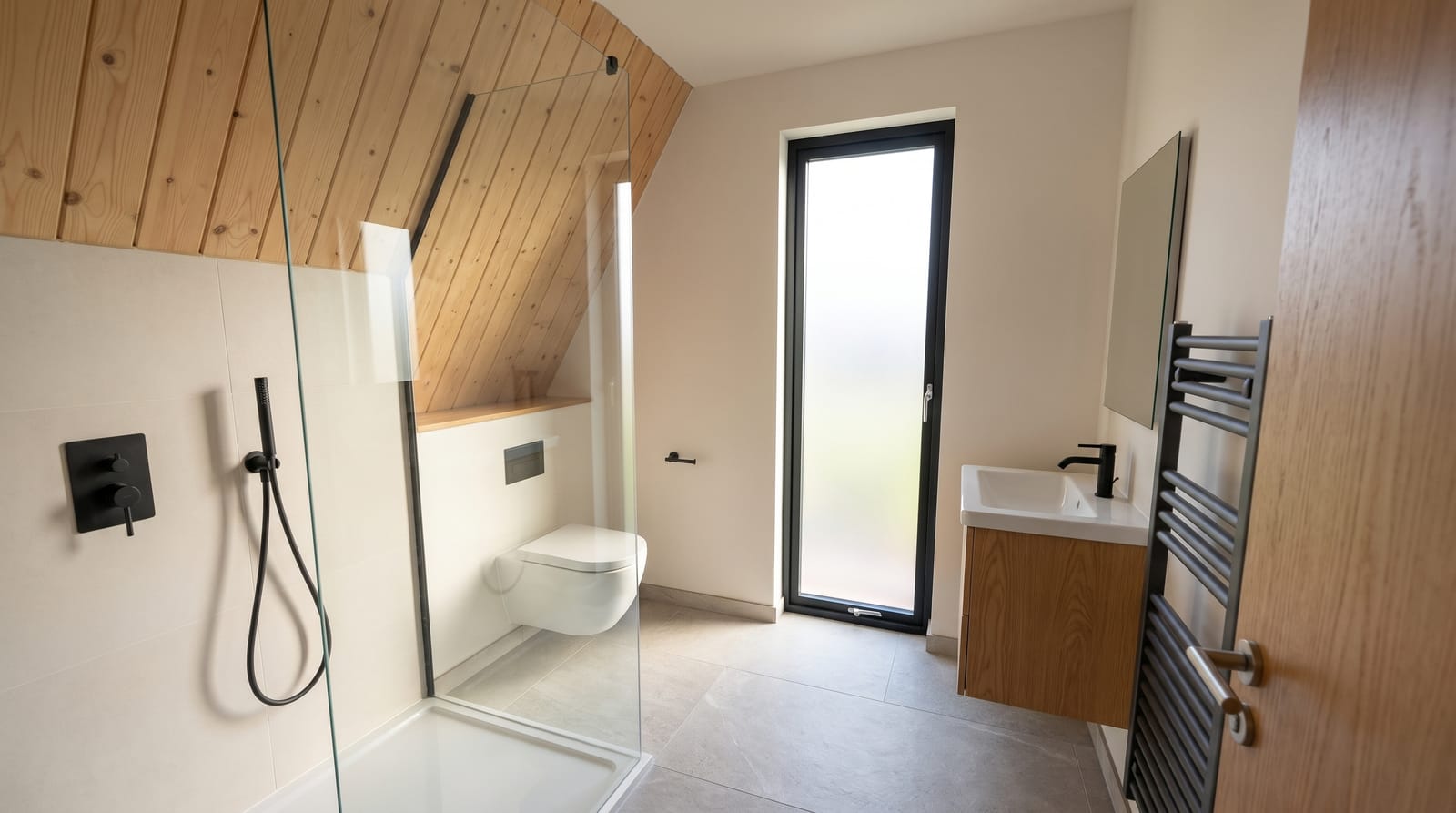 Bathroom under the sloped pine ceiling: walk in shower behind a glass screen, wall hung toilet, oak vanity, black fittings and a tall frosted window