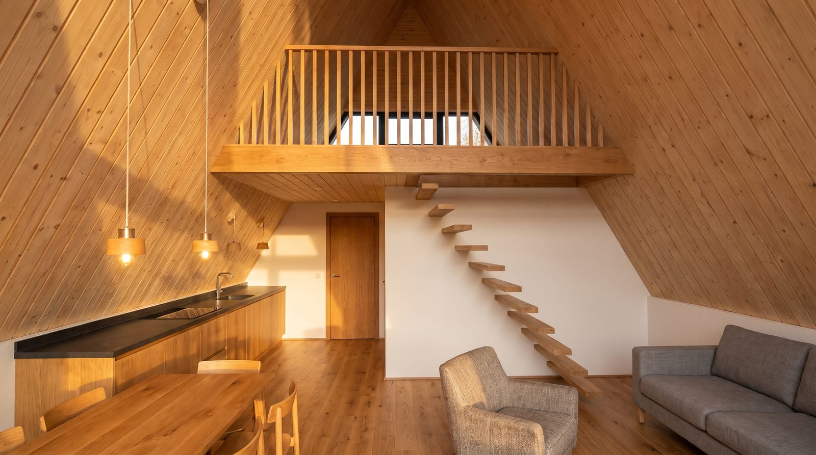 Open living room and kitchen inside the A frame: oak kitchen with a black worktop, dining table, grey sofa and armchair, floating oak stairs up to the loft