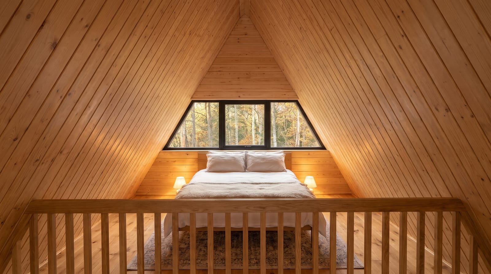 Loft bedroom under the A frame roof: double bed between two bedside lamps, pine clad sloped walls, a wide window to the forest and a timber railing